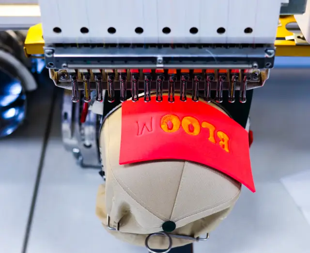 Embroidery machine stitching orange 3D puff lettering through a red foam sheet on a tan cap held in a cap frame