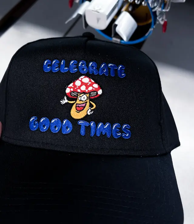 Black structured cap with multi-color flat embroidery of a cartoon mushroom and blue bubble letters, machine behind