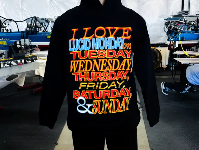 Black hoodie on a form with a large multi-color list of weekdays printed across the chest and kangaroo pocket, presses behind