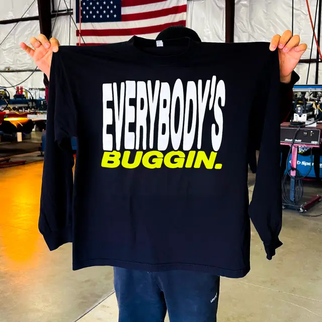 Person holds up a black long sleeve tee with a white and neon yellow text print, with screen printing presses behind