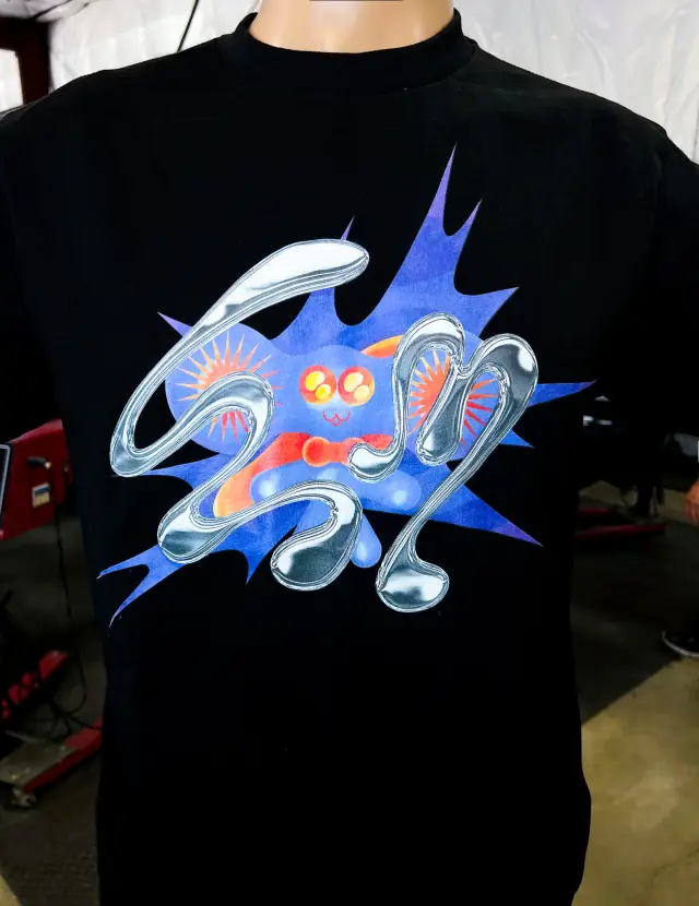 Black tee on a form with a front print of chrome-effect lettering over a blue starburst with a cute creature