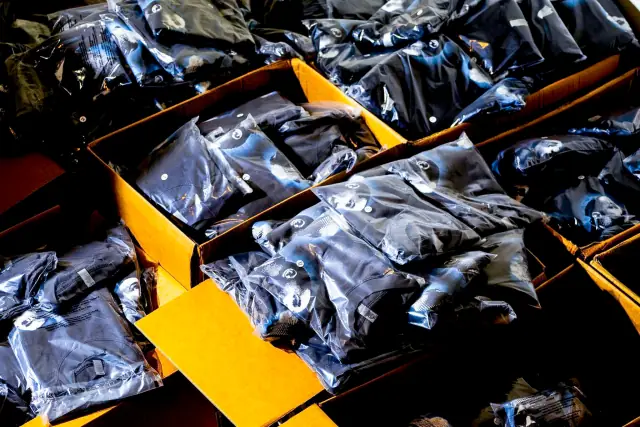 Folded black garments in clear polybags with round size stickers, packed into open cardboard shipping boxes