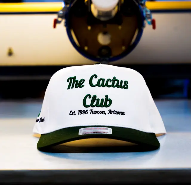 White structured cap with a dark green brim and green and black script embroidery on the front, a machine blurred behind