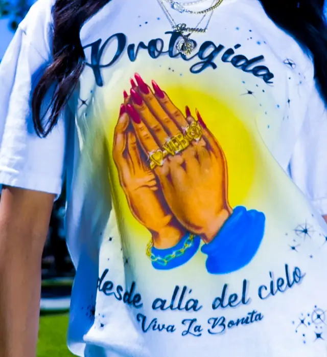 Woman wearing a white tee with an airbrush-style print of praying hands with red nails and gold rings, outdoors